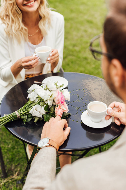 cropped image of couple in autumn outfit sitting at table in cafe with cups of coffee and bouquet on table - Photo, Image