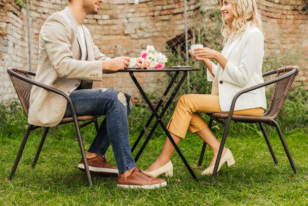 боковой вид на пару в осеннем наряде, сидящую за столом в кафе с чашками кофе и букетом на столе
 - Фото, изображение