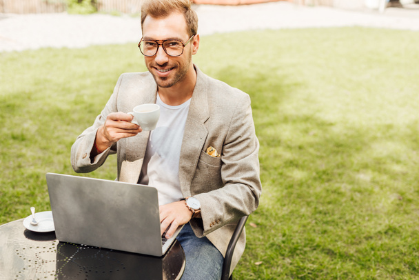 sonriente freelancer en traje de otoño trabajando con el ordenador portátil en la mesa en la ciudad y sosteniendo la taza de café
 - Foto, imagen