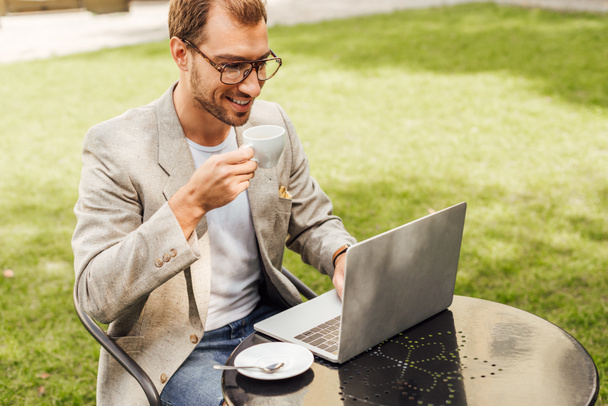 handsome freelancer in autumn outfit working with laptop at table in city and holding cup of coffee - Photo, Image