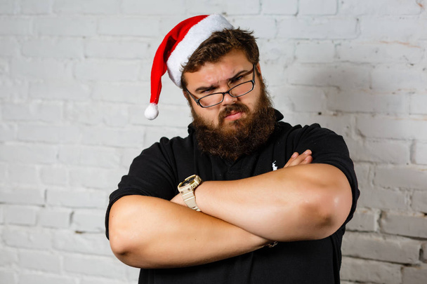 Triste jeune homme caucasien vêtu d'un t-shirt décontracté et d'un chapeau rouge du Père Noël avec de la boue blanche en fourrure, fatigué et ennuyé, homme barbu au chapeau de Père Noël
 - Photo, image