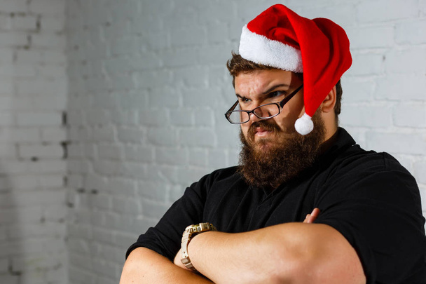Triste jeune homme caucasien vêtu d'un t-shirt décontracté et d'un chapeau rouge du Père Noël avec de la boue blanche en fourrure, fatigué et ennuyé, homme barbu au chapeau de Père Noël
 - Photo, image