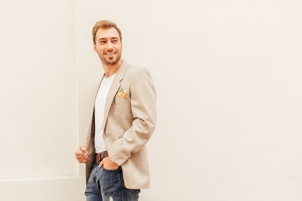 smiling handsome man in jacket and jeans standing near beige wall in city - Foto, afbeelding