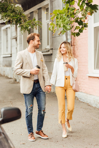 wesoły para w jesienny strój, trzymając się za ręce, patrząc na siebie i chodzenie po ulicy z filiżanki kawy jednorazowe - Zdjęcie, obraz