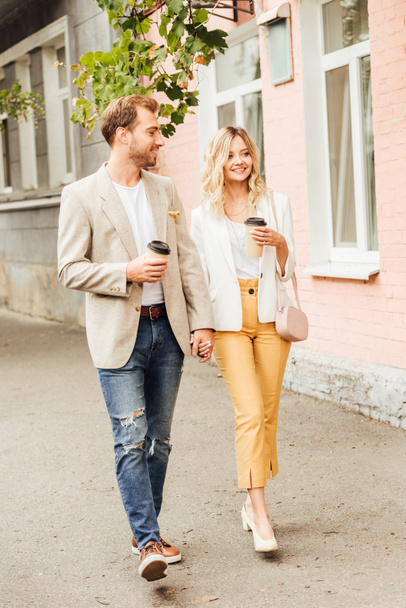 smiling couple in autumn outfit holding hands and walking on street with disposable coffee cups - Фото, изображение