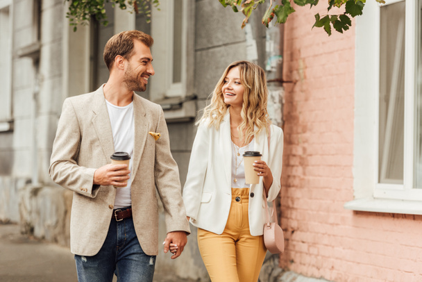 smiling couple in autumn outfit holding hands, looking at each other and walking on street with disposable coffee cups - Photo, Image