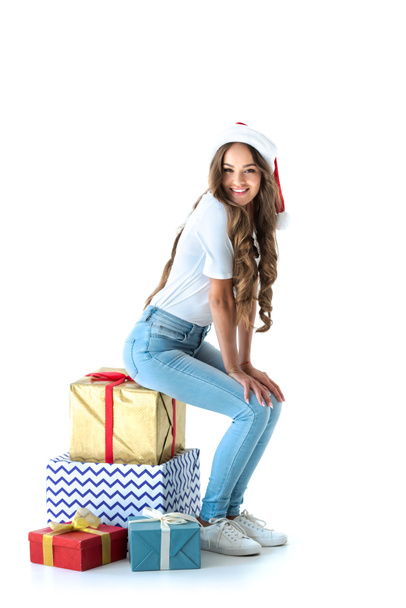 beautiful smiling girl in santa hat sitting on christmas presents, isolated on white - 写真・画像