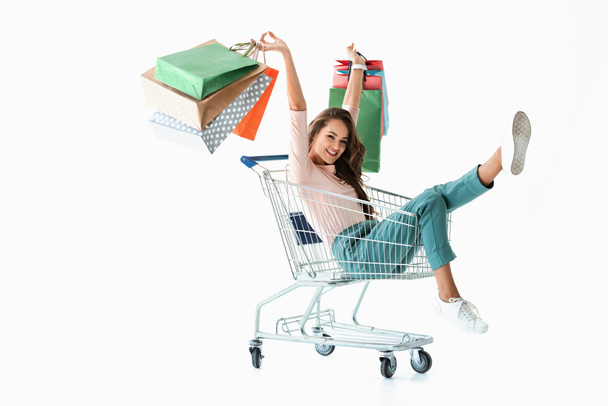 excited girl with shopping bags sitting in shopping cart, isolated on white - Photo, Image