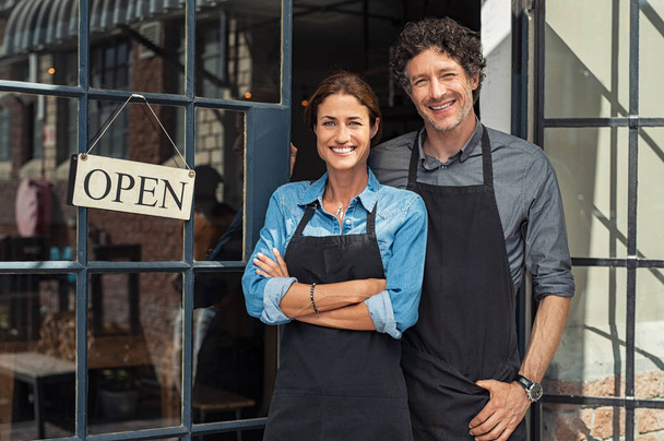 Two cheerful small business owners smiling and looking at camera while standing at entrance door. Happy mature man and mid woman at entrance of newly opened restaurant with open sign board. Smiling couple welcoming customers to small business shop. - Φωτογραφία, εικόνα