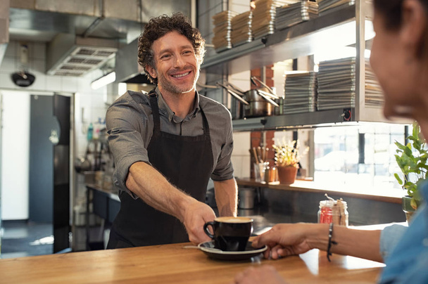 Smiling mature waiter giving hot coffee to woman at cafeteria. Happy man wearing black apron standing behind counter giving a cappuccino cup to woman in a coffee shop. Portrait of cheerful man serving happy customer at restaurant. - Foto, Bild