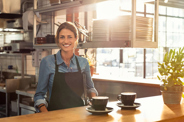 Smiling waitress wearing black apron standing behind counter in cafeteria and looking at camera. Mature woman serving two cups of hot coffee to customer in cafeteria. Small business and entrepreneur concept. - Φωτογραφία, εικόνα