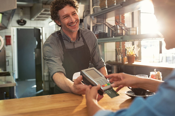 Woman paying by credit card and entering pin code on reader holded by smiling barista in cafeteria. Customer using credit card for payment. Mature cashier wearing apron accepting payment over nfc technology. - Photo, Image