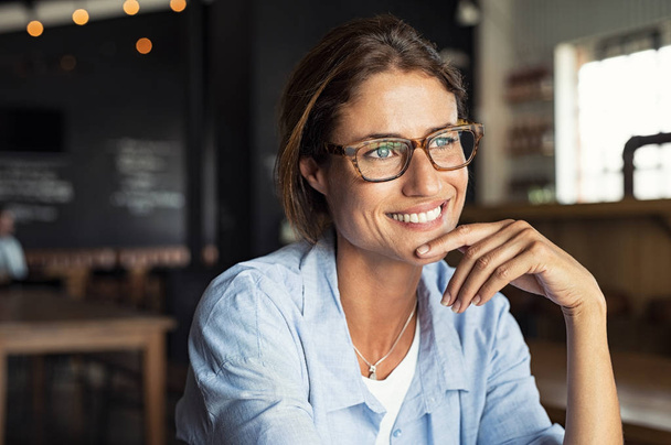 Portrait of beautiful mature woman sitting in cafeteria looking away. Cheerful mature woman wearing eyeglasses thinking with finger on chin. Happy woman relaxing at cafe and smiling. - Foto, Bild