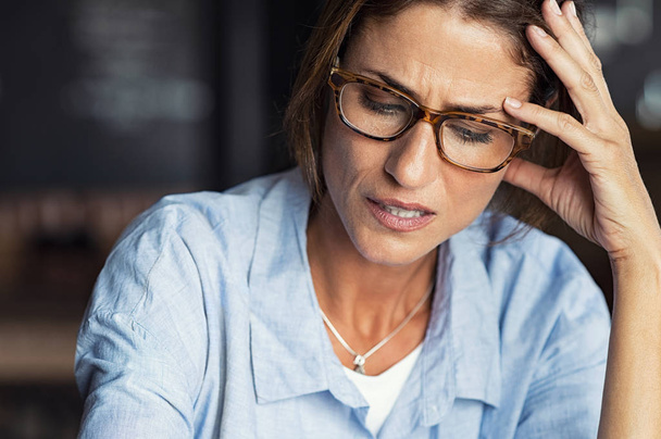 Portrait of stressed mature woman with hand on head looking down. Worried woman wearing spectacles. Tired lady having headache sitting indoors. - Foto, Bild