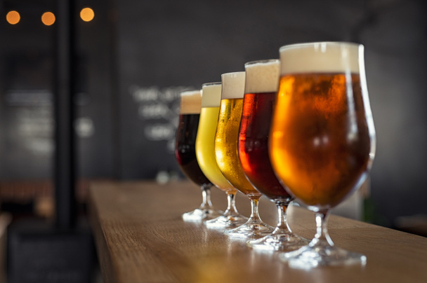 Glasses with different sorts of craft beer on wooden bar. Tap beer in pint glasses arranged in a row. Closeup of five glasses of different types of draught beer in a pub. - 写真・画像