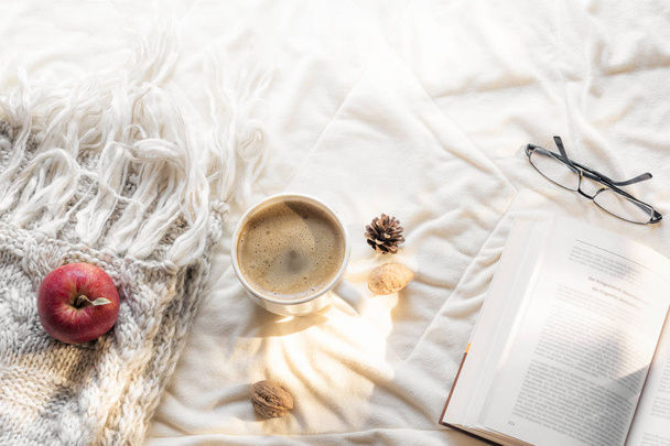 Cozy leisure concept with an open book, glasses, an apple on a fluffy shawl and a hot cup of coffee, on a white blanket, in the morning sun. - Photo, Image