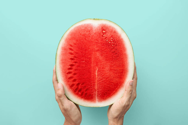 Handen met een helft van rode watermeloen op een blauwe achtergrond, bovenaanzicht met uitknippad - Foto, afbeelding