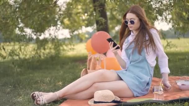 Ginger girl in glasses and jeans skirt making selfie during picnic in park - 映像、動画