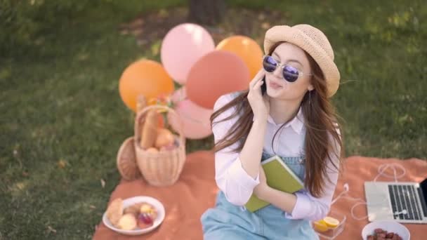 Cheerful ginger college student in straw hat and sunglasses talking on phone in park - Záběry, video