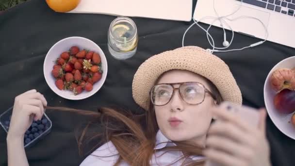 Red haired young woman making funny faces and selfies lying in park - Materiaali, video