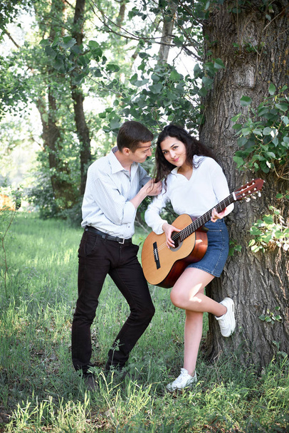 young couple walking in the forest and playing guitar, summer nature, bright sunlight, shadows and green leaves, romantic feelings - Valokuva, kuva