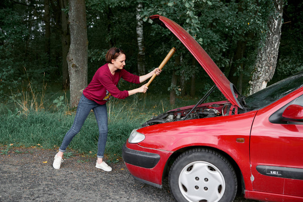 μια νεαρή γυναίκα βρίσκεται κοντά σε ένα σπασμένο αυτοκίνητο, έχει άγχος και οργή, θέλει να συνθλίψει το αυτοκίνητό της με ένα ρόπαλο του μπέιζμπολ - Φωτογραφία, εικόνα