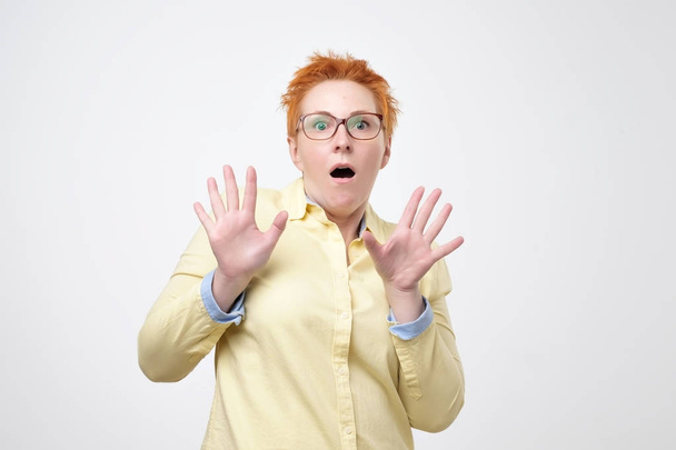 Oh no concept. Headshot of attractive european woman with glasses dressed in yellow shirt looking at camera, keeping mouth wide opened feeling shocked and stressed. - Foto, imagen