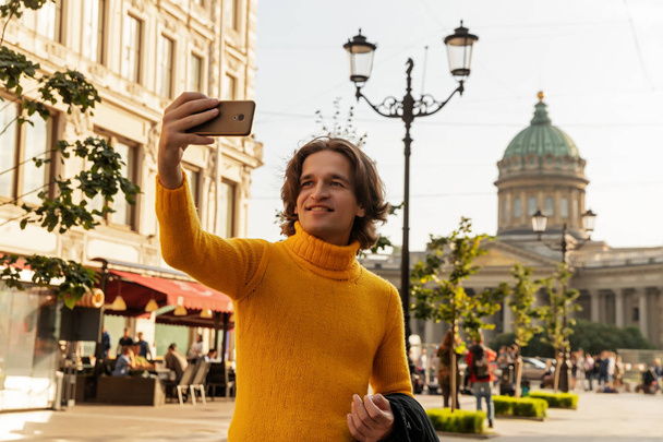 Le jeune homme attend quelqu'un et fait du selfie, il s'habille d'un pull jaune, d'un imperméable ou d'une veste noire, d'un jean, d'une rue et d'une cathédrale Kazanskiy sur fond, journée ensoleillée
 - Photo, image