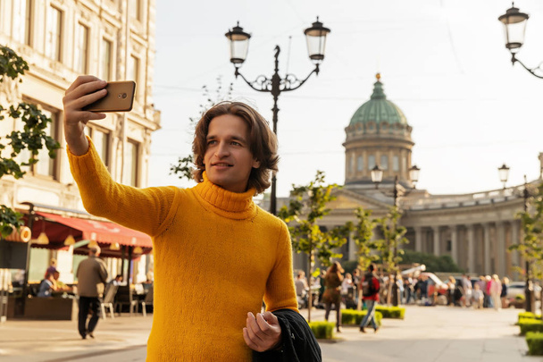 The young man waiting someone and does selfie, he dressed in a yellow sweater, a black raincoat or jacket, jeans, street and Kazanskiy cathedral on background, sunny day - Photo, image