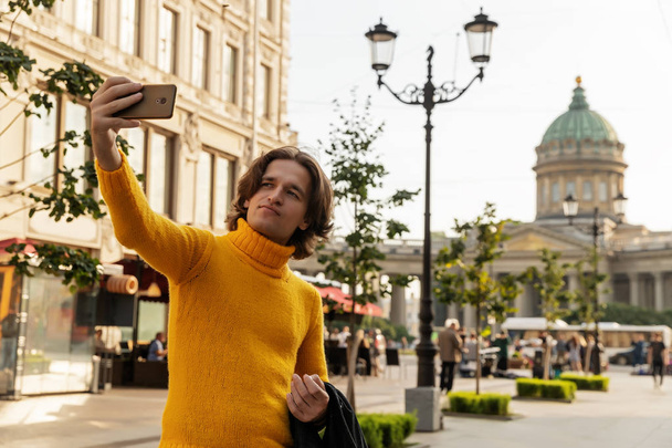 The young man waiting someone and does selfie, he dressed in a yellow sweater, a black raincoat or jacket, jeans, street and Kazanskiy cathedral on background, sunny day - Fotoğraf, Görsel