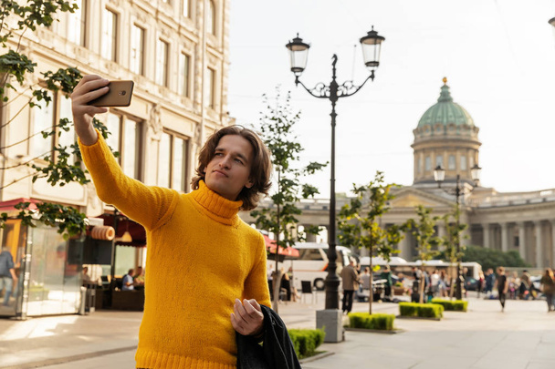 The young man waiting someone and does selfie, he dressed in a yellow sweater, a black raincoat or jacket, jeans, street and Kazanskiy cathedral on background, sunny day - Φωτογραφία, εικόνα