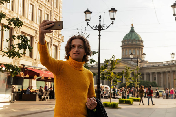 The young man waiting someone and does selfie, he dressed in a yellow sweater, a black raincoat or jacket, jeans, street and Kazanskiy cathedral on background, sunny day - Φωτογραφία, εικόνα