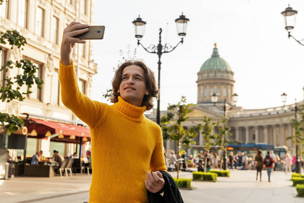 El joven esperando a alguien y hace selfie, se vistió con un suéter amarillo, un impermeable negro o chaqueta, jeans, calle y catedral de Kazanskiy en el fondo, día soleado
 - Foto, Imagen
