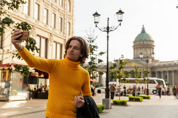 Ο νεαρός άντρας περιμένει κάποιος και κάνει selfie, αυτός ντυμένος με ένα κίτρινο πουλόβερ, ένα μαύρο αδιάβροχο ή μπουφάν, τζιν, Οδός και Καθεδρικός Ναός Kazanskiy σε φόντο, ηλιόλουστη μέρα - Φωτογραφία, εικόνα