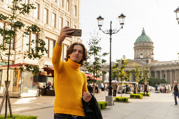 The young man waiting someone and does selfie, he dressed in a yellow sweater, a black raincoat or jacket, jeans, street and Kazanskiy cathedral on background, sunny day - 写真・画像