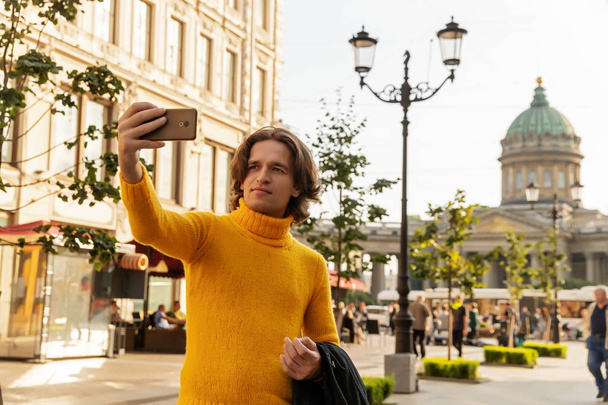 The young man waiting someone and does selfie, he dressed in a yellow sweater, a black raincoat or jacket, jeans, street and Kazanskiy cathedral on background, sunny day - Foto, Imagen
