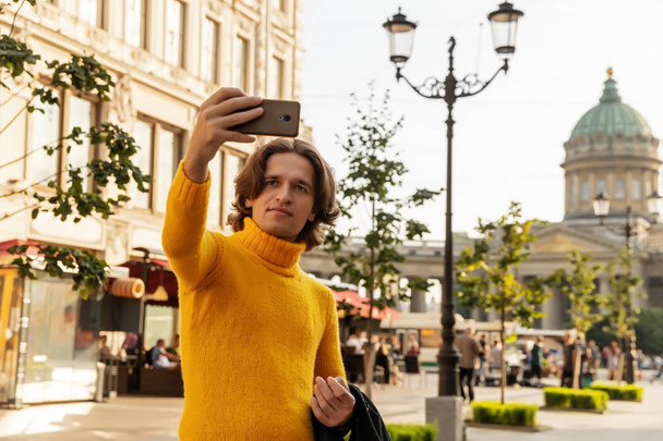 The young man waiting someone and does selfie, he dressed in a yellow sweater, a black raincoat or jacket, jeans, street and Kazanskiy cathedral on background, sunny day - Фото, изображение