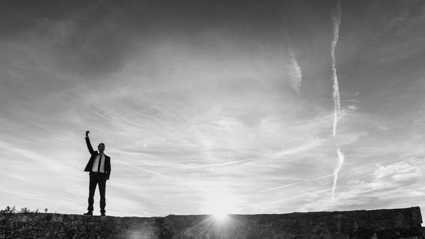 Ликующий успешный бизнесмен бьет кулаком по воздуху, стоя на стене, подсвеченной солнечным вспышкой от заходящего солнца в концептуальном черно-белом изображении
. - Фото, изображение