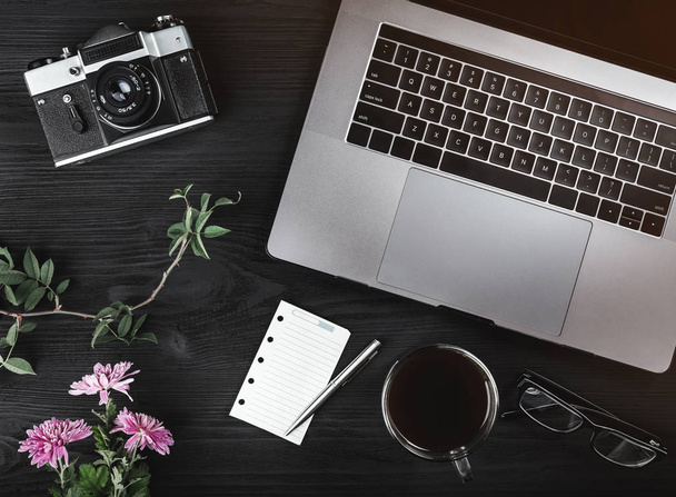 Top view of notebook computer, eyeglasses, cup of coffee and white sheet, vintage camera and pink flowers over black stone background - Photo, Image