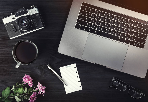 Cup of tea or coffee, photo camera, laptop computer, eyeglasses and pink flowers on black wooden table. Top view, flat lay - Fotografie, Obrázek