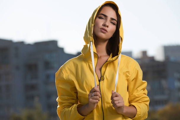 Confident woman outdoor portrait at city background in yellow windbreaker under hood - Zdjęcie, obraz