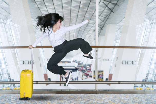 Happy businesswoman jumping in the airport terminal while holding a luggage - Foto, afbeelding