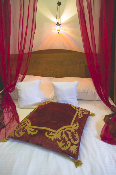 赤い透明なカーテンと赤と金色のクッションが付いている快適な天蓋のベッド - 写真・画像