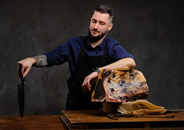 Chef cocinero sostiene cuchillo y posando cerca de una mesa con carne seca exclusiva sobre un fondo oscuro
. - Foto, Imagen