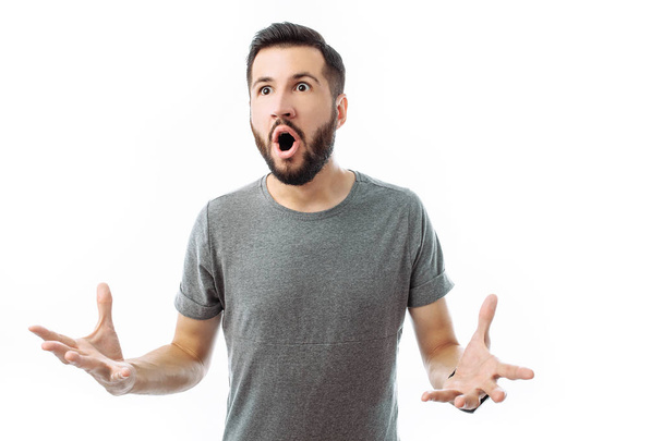 Portrait of surprised and delighted man with beard, in grey t-shirt, standing with hands up and looking at camera isolated on white background - Photo, image
