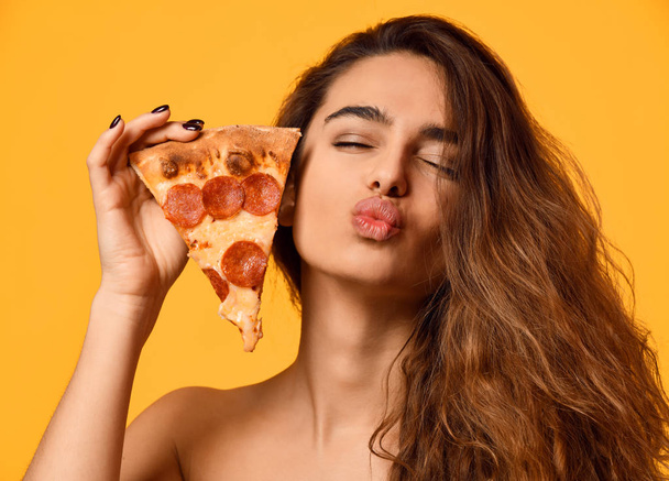 Young beautiful woman eat hold pepperoni pizza slice show kissing sign with lips on yellow - Photo, Image