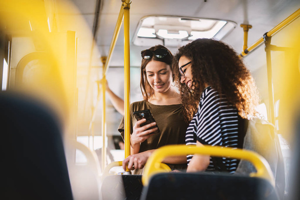 two cheerful pretty young women standing in a bus and looking at phone  - Foto, Imagem