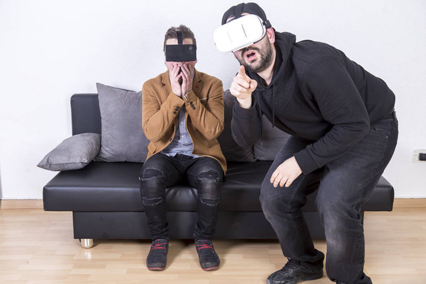 two amazed and shocked men using VR goggles and sitting on sofa in room  - Foto, Bild