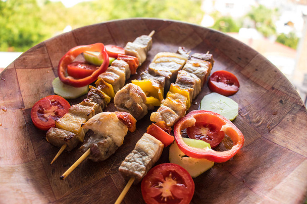 Skewered meat pieces prepared on grill with vegetables. Barbecued shish kebab or shashlik on sticks. Outdoor picnic meal on wooden round dish - Photo, Image