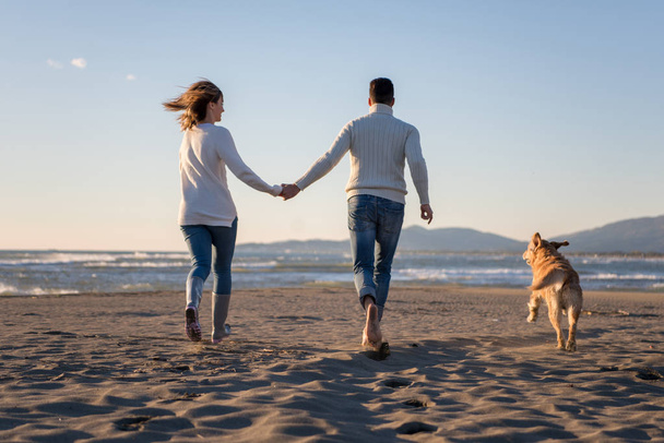 Casal correndo na praia segurando as mãos com o cão no dia de outono - Foto, Imagem